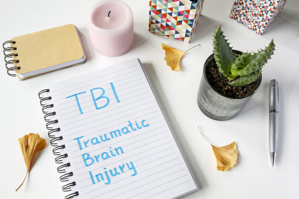 The words “TBI Traumatic Brain Injury” written in a notebook on a white table with a pink candle, pen, and plant