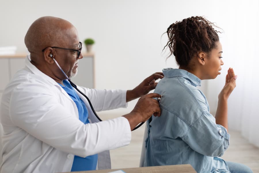 Doctor listening to coughing female patient’s lungs with stethoscope