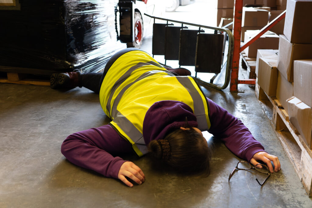 Woman in yellow safety vest lying warehouse floor after falling from ladder