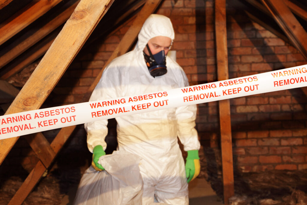 A man wearing protective clothing and respirator with a warning asbestos removal banner in front of him