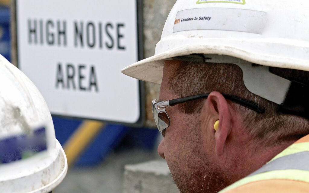 Close up of a construction worker wearing earplugs, protective glasses and hard hat with a high noise area sign in the background.