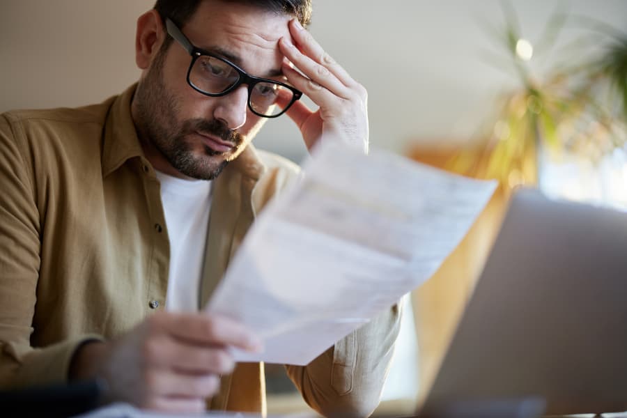 A man with glasses and a beard in a tan shirt holding his head while looking at a paper