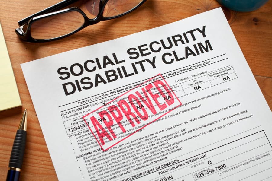 Close up of a Social Security Disability claim with a red approved stamp on it and a pen and eyeglasses on a wooded desk