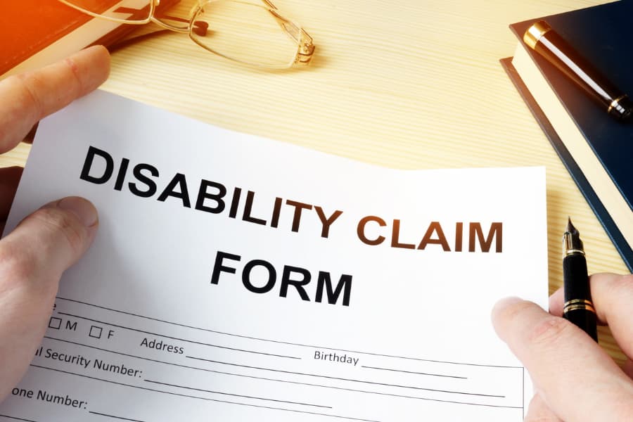 Close-up of a man holding a disability claim form and pen with eyeglasses and a book on a wood desk