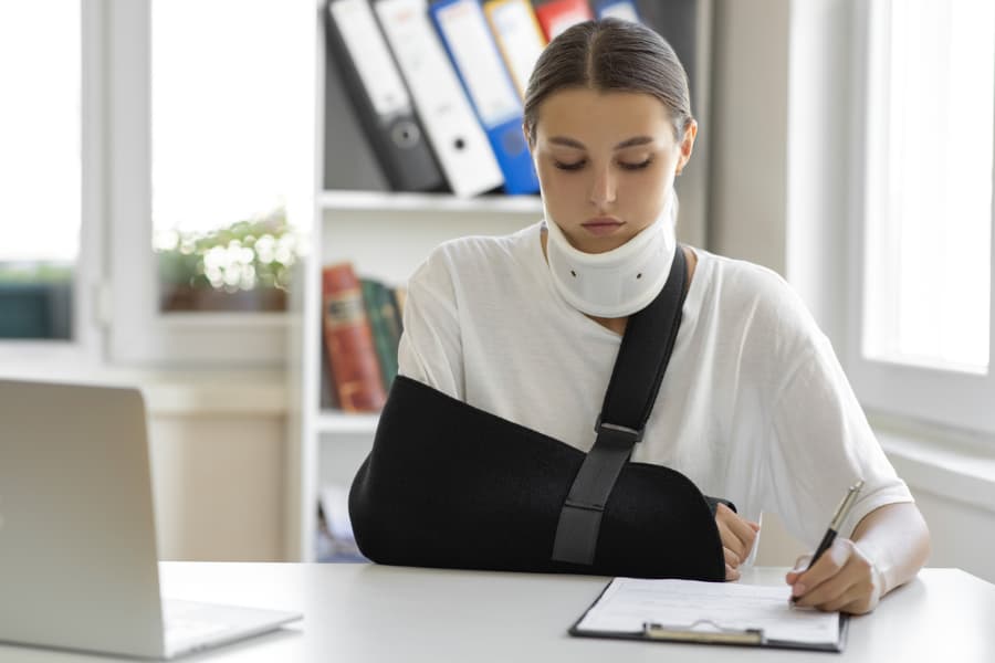 Woman with a neck brace and arm in sling sitting at a desk filling out an SSDI form